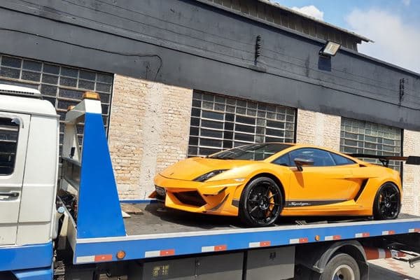 Carro esportivo laranja carregado sobre a plataforma azul do guincho ME, em frente a um galpão de tijolo.